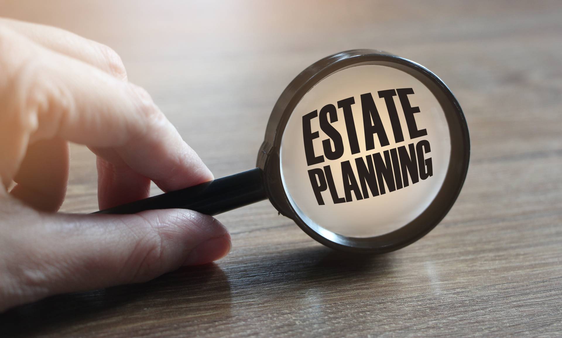 A hand holding a small magnifying glass that is enhancing the text "estate planning" on a wooden table.