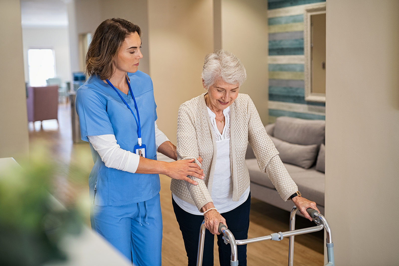 A nurse helping an older woman who is using a walker move across a room of the nursing home.
