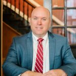 Headshot of attorney & owner of Crow Estate Planning & Probate, John Crow sitting in front of a wooden glass door in a suit.