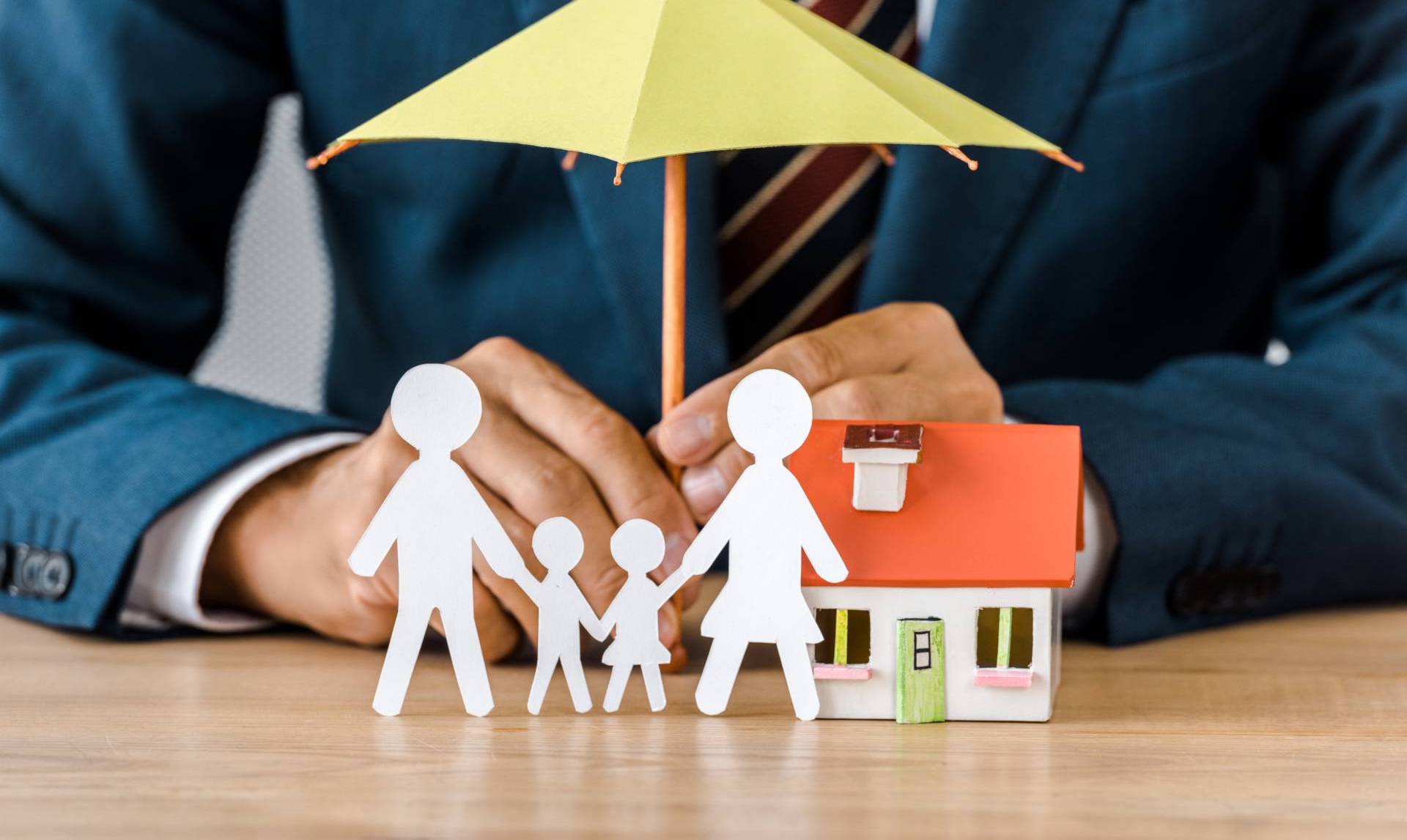 A person in a suit putting together a small representation of a family in front of a house with an umbrella covering them.