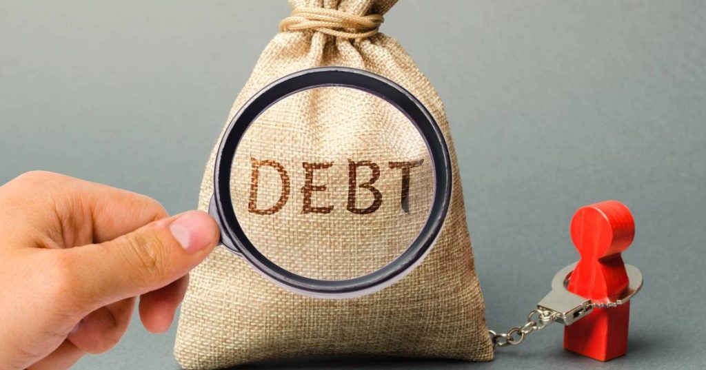 A hand has a magnifying glass over a money bag that has the text "debt" on it and is cuffed to a wooden red toy person.