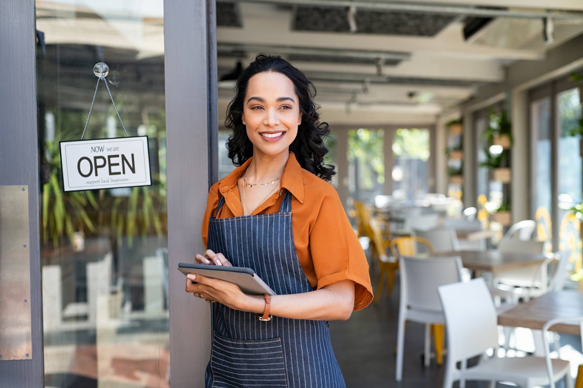 A business person standing in front of their cafe that is open, excited for customers to come inside.