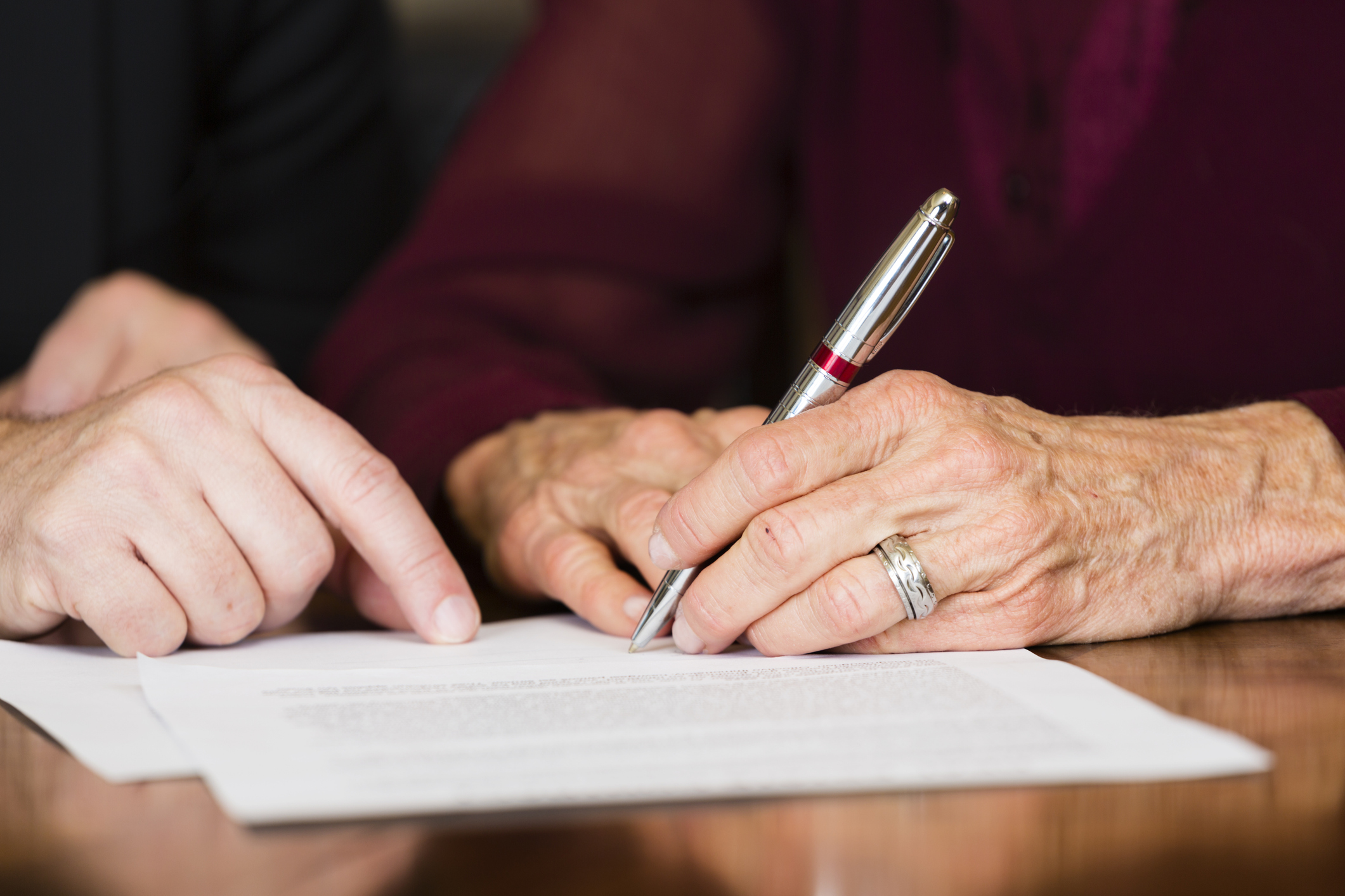 The hands of an older man signing a contract related to his estate plan as a lawyer shows important parts of the document.