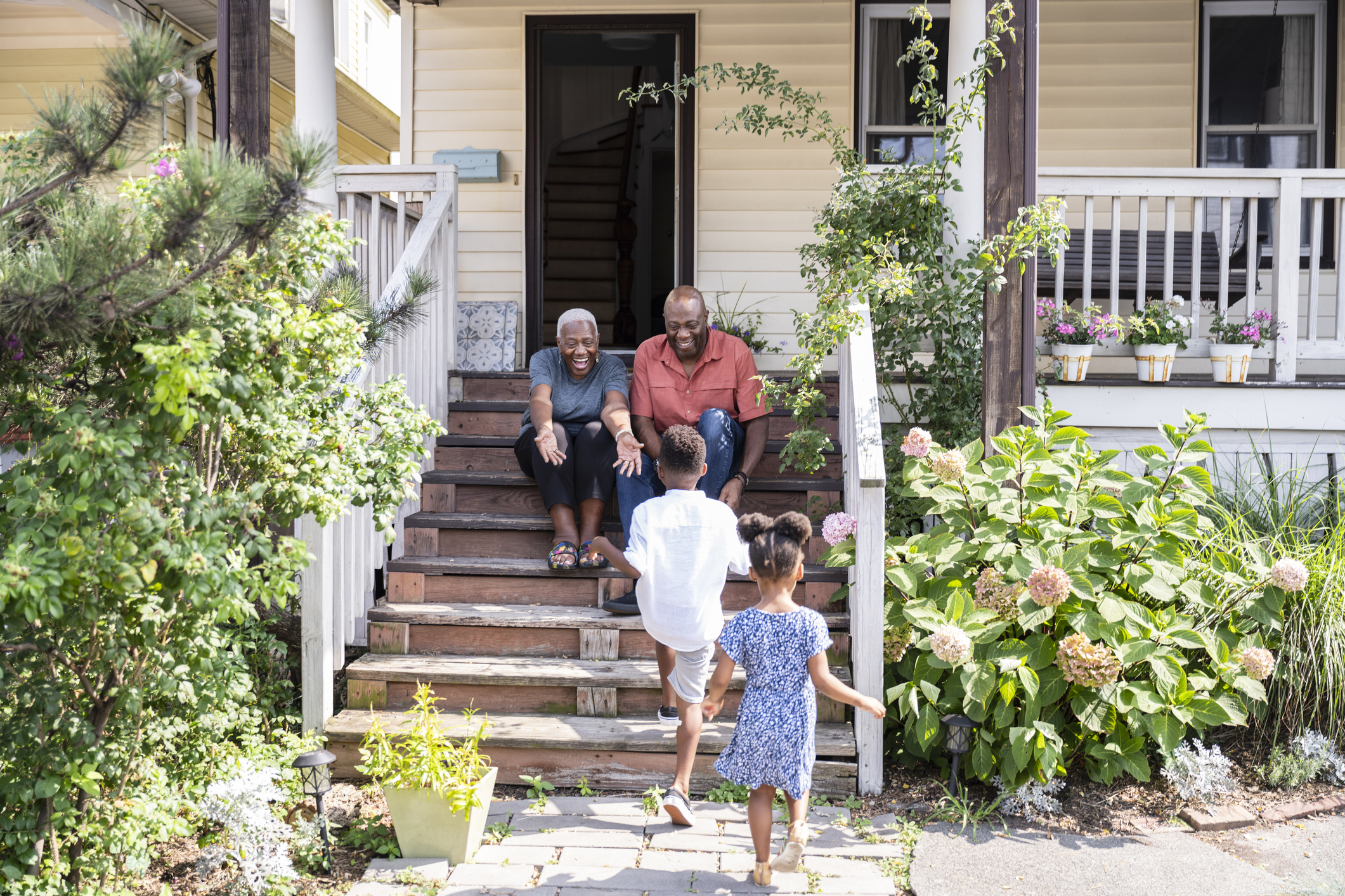 Two siblings walking up to their elderly grandparents who are sitting on the steps of their front porch, excited to see them.