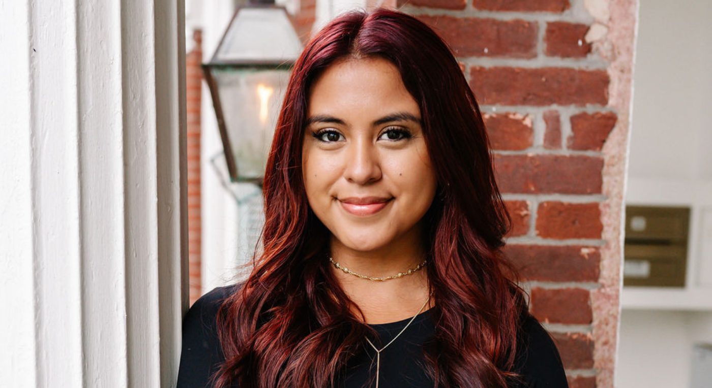 A professional headshot of a woman with vibrant dark red hair, smiling softly in front of a brick wall and white pillar.