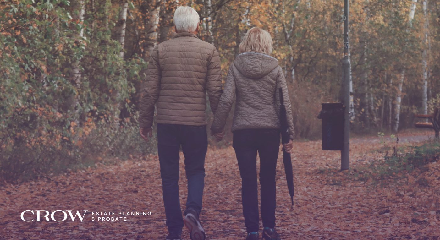 An elderly couple walks hand-in-hand down a leaf-strewn path in a park during autumn.